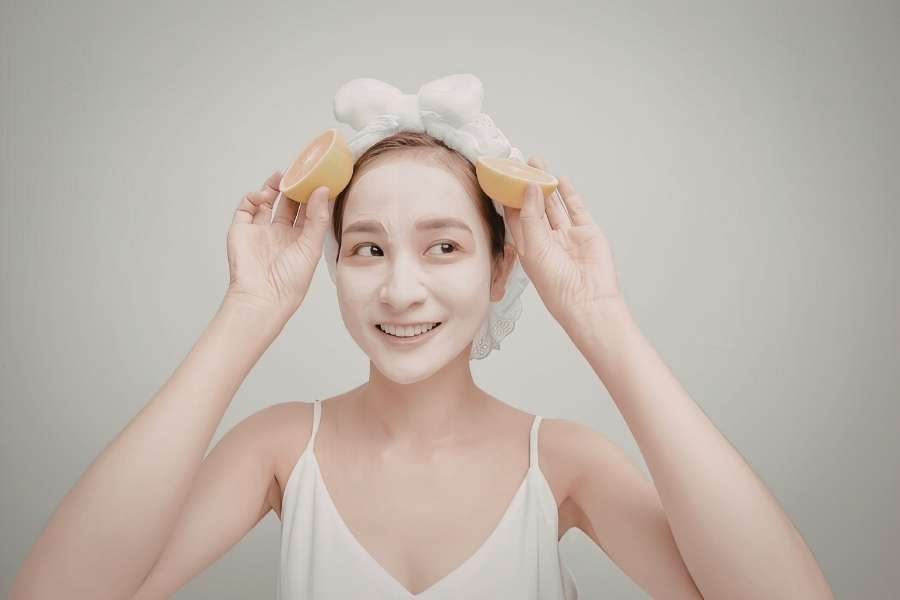 Jeune femme souriante avec un masque blanc sur le visage tenant deux tranches de citron, illustrant des soins du visage maison aux ingrédients naturels.
