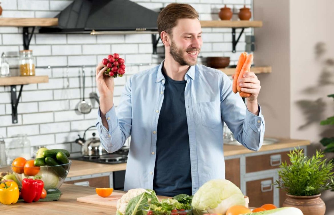 Un homme souriant dans une cuisine tenant des radis et des carottes, illustrant la préparation d'un repas sèche homme équilibré.