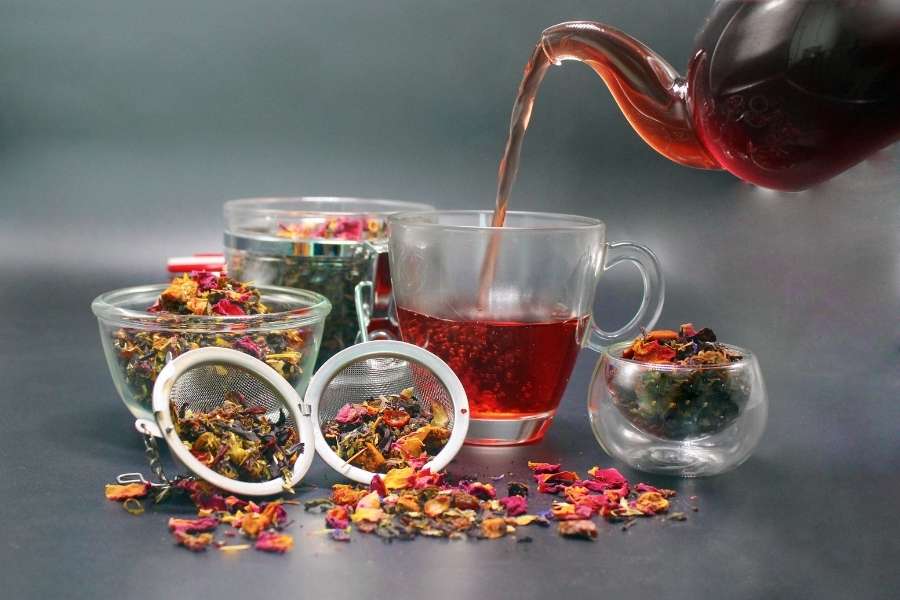 Une théière verse une infusion rougeoyante dans une tasse transparente entourée de mélanges de plantes pour tisanes puissantes.