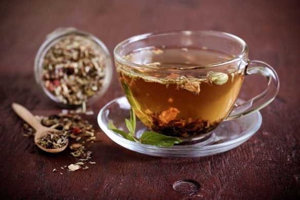 Une tasse transparente remplie d'une infusion chaude et de plantes pose sur une soucoupe en bois aux côtés de tisanes puissantes.